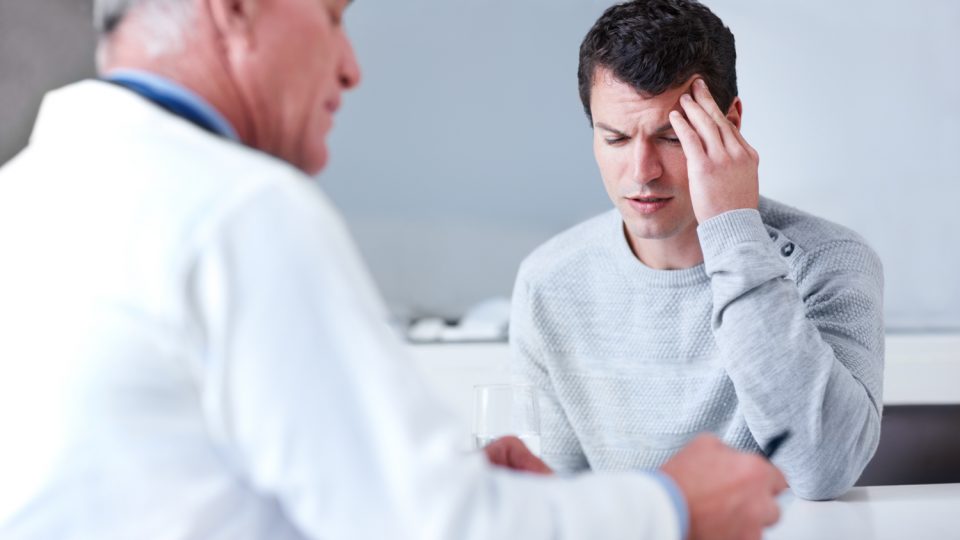 Patient massaging the side of their head with their fingers, seated across from a doctor taking notes