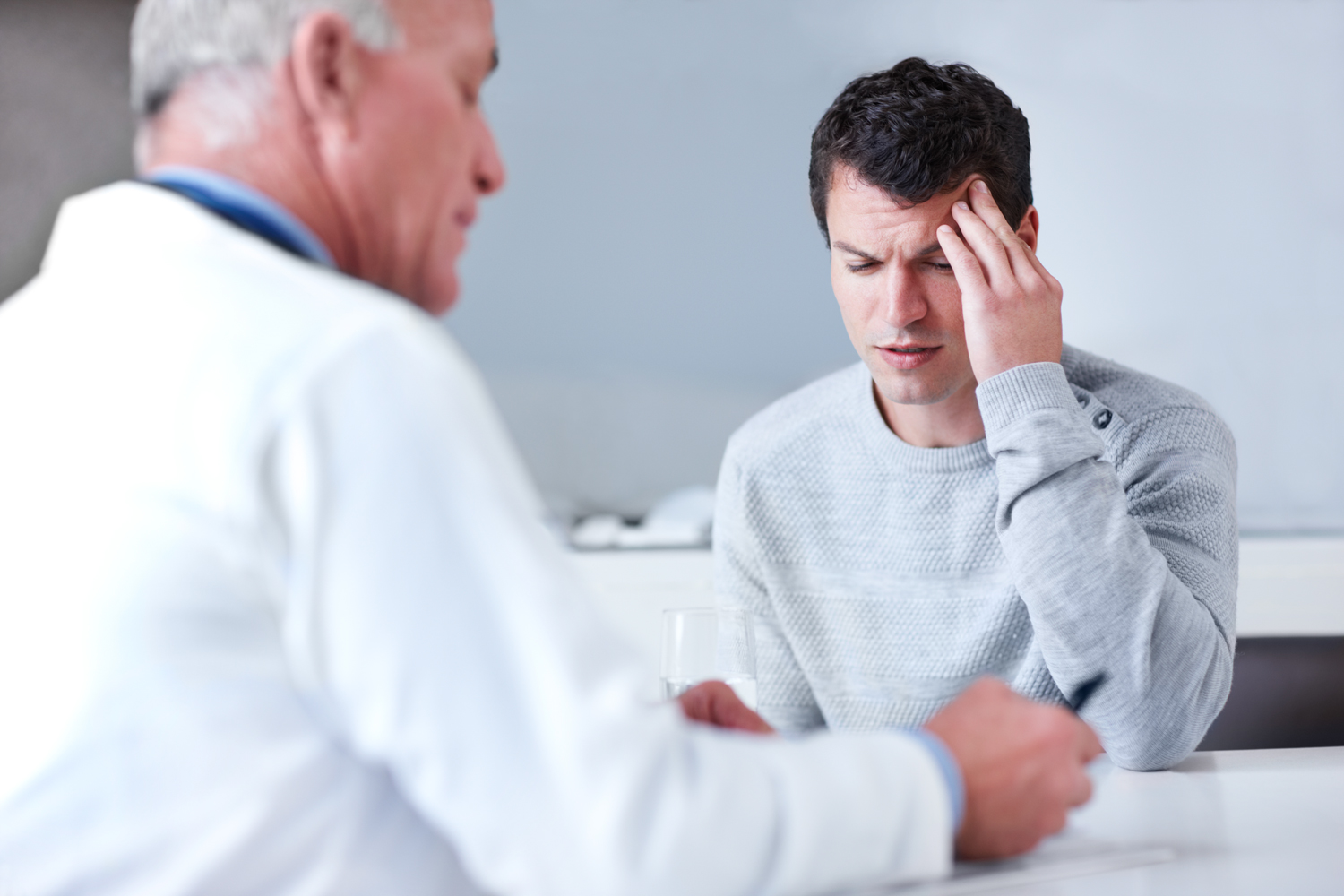 Patient massaging the side of their head with their fingers, seated across from a doctor taking notes